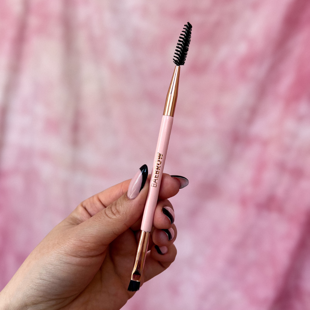 Brow styling brush with pink handle and rose gold accents held by a hand against a pink background