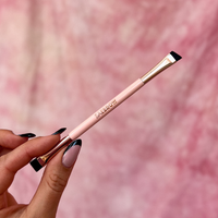 Brow styling brush with pink handle and rose gold accents held by a hand against a pink background