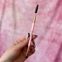 Brow styling brush with pink handle and rose gold accents held by a hand against a pink background