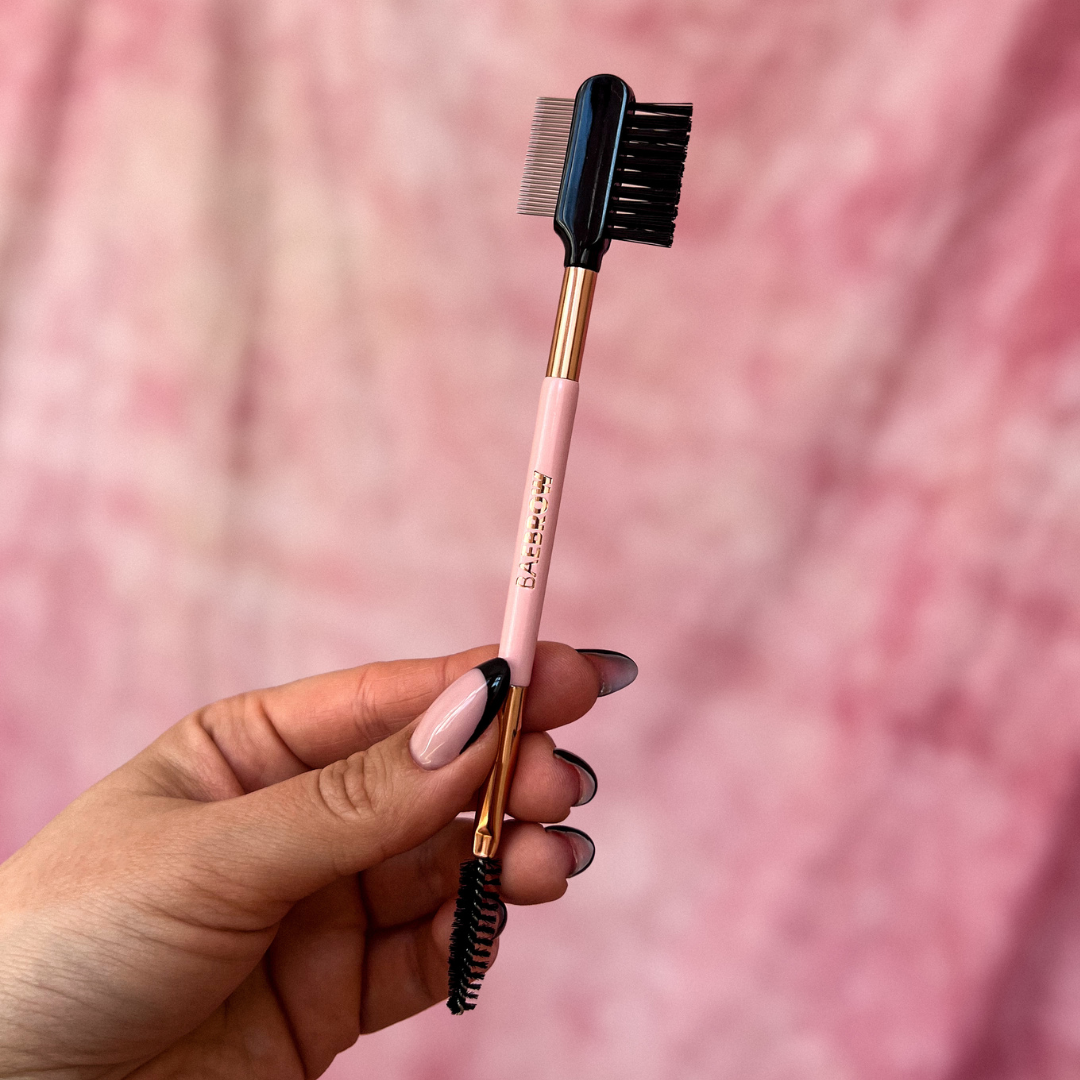 Brow styling brush with pink handle and rose gold accents held by a hand against a pink background